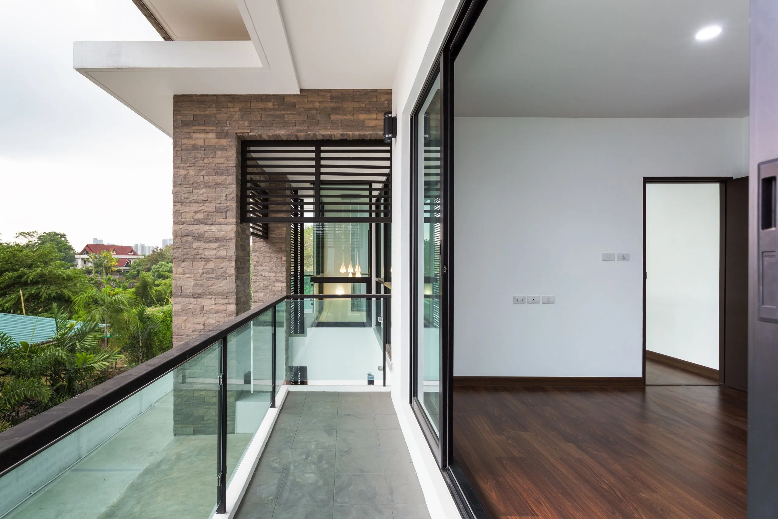 shutterstock_416008192-jpg Modern apartment balcony with glass railing, adjoining room with wooden floor, and view of trees in the background.